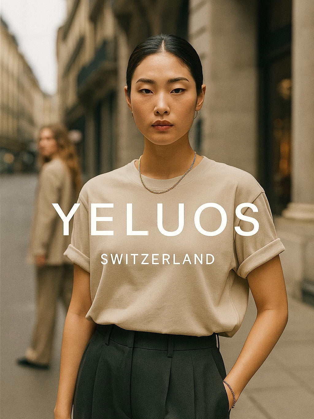 young woman wearing a beige YELUOS Switzerland t-shirt and dark trousers, standing confidently on a European city street with historic stone buildings in the background.